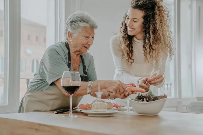 A caregiver and an older woman preparing a meal together in a sunny kitchen